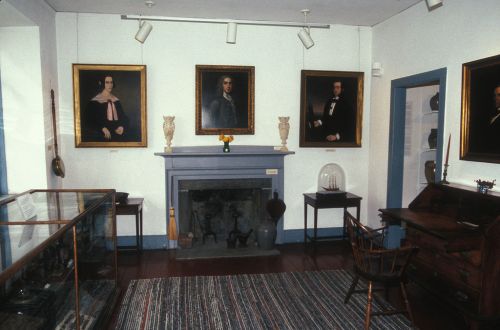 A room with portraits on the walls, a fireplace, a desk, and display cases, featuring a cozy, historical atmosphere.