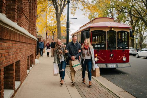 Three friends walk arm in arm on a sidewalk, smiling, with a red trolley and autumn trees in the background.