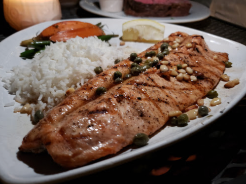 Grilled salmon fillets with capers and pine nuts, served with rice and a slice of lemon on a white plate.