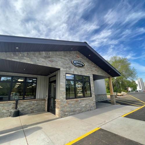 Exterior view of a stone and metal building with a sign reading "Mullins," surrounded by trees and a clear blue sky.
