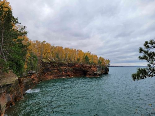 A rocky shoreline with vibrant autumn trees along the edge, overlooking a calm lake under a cloudy sky.