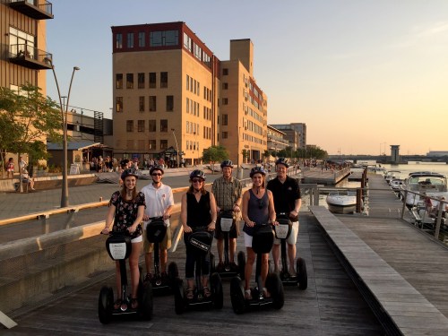 A group of six people on Segways, smiling, with a waterfront and buildings in the background during sunset.