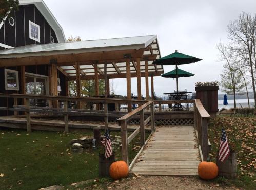 A wooden walkway leads to a lakeside building with a patio, umbrellas, and pumpkins by the entrance.