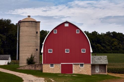 A red barn with a silo beside it, set against a backdrop of green fields and a cloudy sky.
