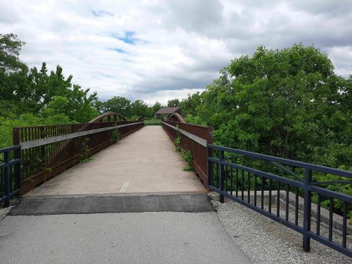 A pedestrian bridge surrounded by greenery under a cloudy sky, leading into a lush landscape.