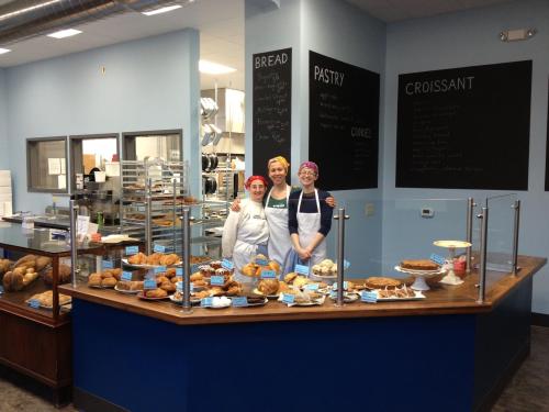 Three bakers stand behind a display counter filled with various baked goods in a bright bakery setting.