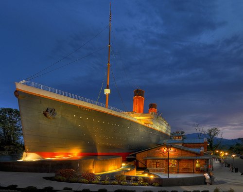 A large ship replica illuminated at night, with a nearby building, set against a cloudy sky.