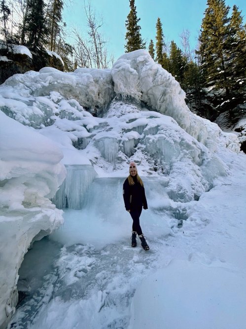 A person stands in front of a frozen waterfall surrounded by snow and tall evergreen trees.