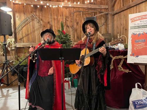Two women in festive attire perform with a guitar in a rustic setting, decorated for the holidays.