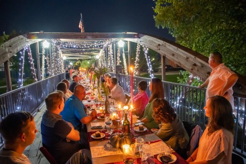 A long table set for dinner under string lights on a bridge, with guests enjoying a festive evening.