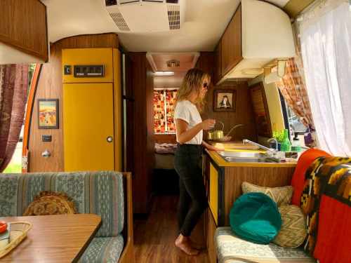 A woman stands in a cozy camper kitchen, cooking with colorful decor and a dining area in the foreground.