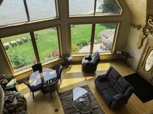 A spacious living room with large windows overlooking a lake and green lawn, featuring a round table and comfortable seating.