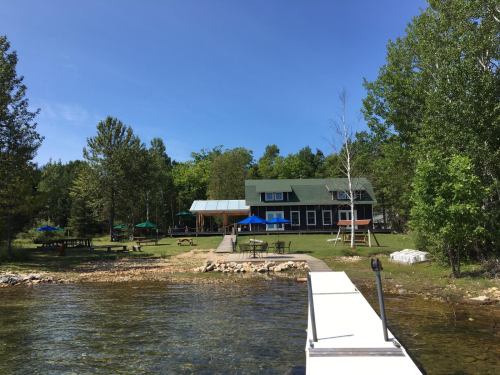 A serene lakeside view featuring a dock, green trees, and a large cabin with outdoor seating areas.