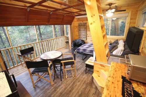 Cozy wooden cabin interior with a dining table, chairs, and a sofa, surrounded by large windows overlooking nature.