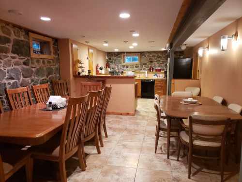Cozy dining area with wooden tables and chairs, stone walls, and a kitchen in the background. Warm lighting throughout.