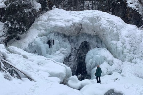 A person stands near a frozen waterfall surrounded by snow-covered rocks and trees in a winter landscape.