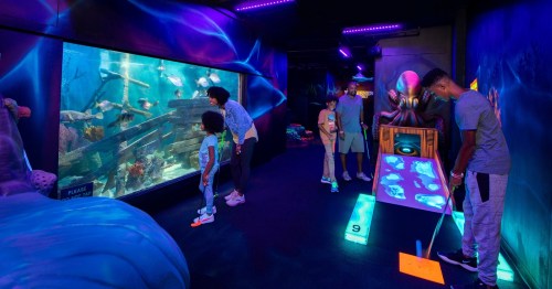 A family enjoys an interactive aquarium exhibit with glowing decorations and a mini-golf setup in a dark room.