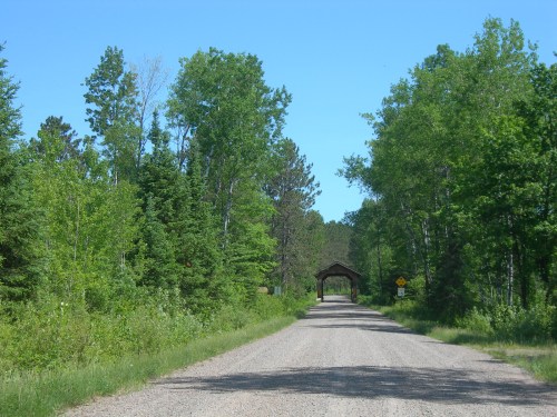 A gravel road lined with lush green trees leads to a covered wooden bridge under a clear blue sky.