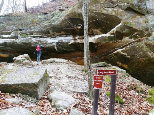 A person stands near a large rock formation with a sign reading "Big Sand Cave" in a wooded area.