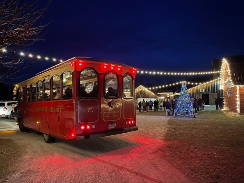 A red trolley bus parked at night, with festive lights and a decorated tree in the background, and people gathered nearby.