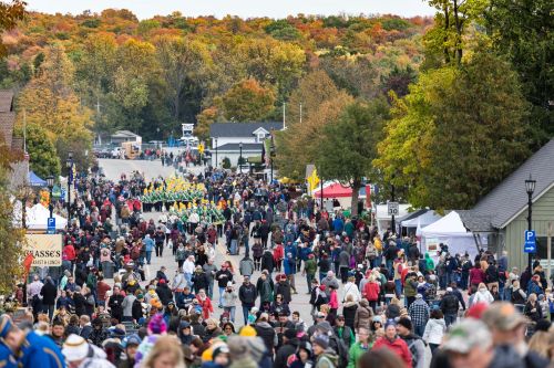 A bustling street filled with crowds and vendors, with a parade marching through colorful autumn trees in the background.