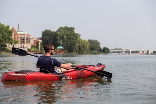 A person kayaking on a calm river, with trees and a building in the background under a clear sky.