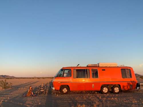 An orange van parked in a desert at sunset, with a person sitting nearby and bicycles beside them.
