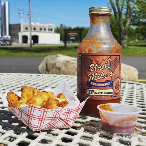 A bottle of Uncle Mike's Maple BBQ sauce next to a basket of crispy fries on a picnic table outdoors.