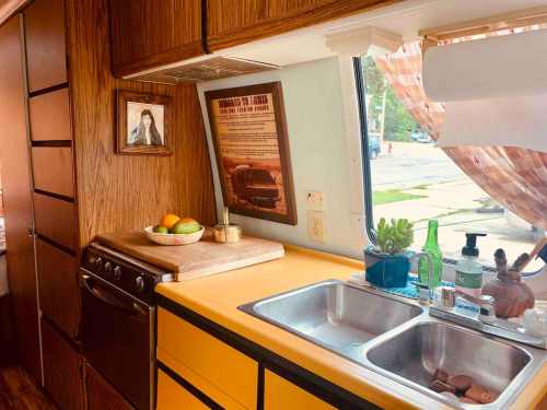 A cozy kitchen with wooden cabinets, a double sink, fruit on a cutting board, and a window with a view.