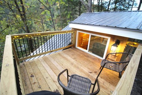 Cozy wooden deck with two chairs, overlooking a forested area, illuminated by soft lights and a lantern.