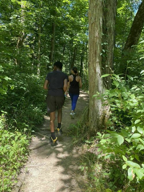 Two people walking on a wooded trail surrounded by lush green trees and plants.