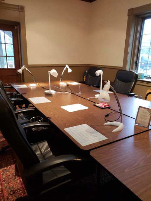 A conference room with a long table, five desk lamps, and black chairs, featuring a window and decorative rug.