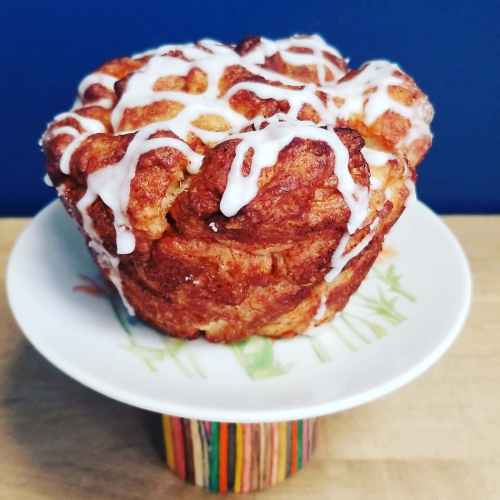 A fluffy cinnamon roll cake drizzled with white icing, displayed on a decorative plate atop a colorful wooden stand.
