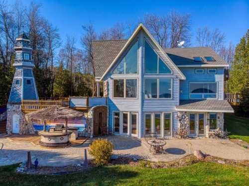 A modern blue house with large windows, a stone patio, and a lighthouse nearby, surrounded by trees and greenery.