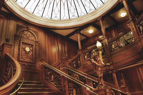 Elegant wooden staircase with ornate railings and a stained glass ceiling, featuring a decorative statue at the landing.