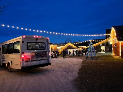 A bus parked near a festive outdoor scene with string lights, a decorated tree, and people gathering at a barn.