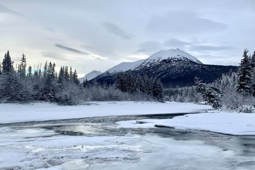 A serene winter landscape featuring a snowy river, frosted trees, and mountains under a cloudy sky.
