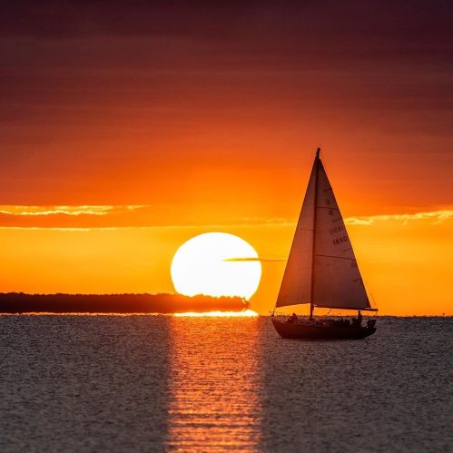 A sailboat glides across the water at sunset, with a large sun setting on the horizon, casting a warm glow.