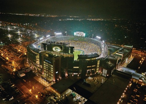 Aerial view of a packed stadium at night, illuminated with lights, surrounded by parking and city lights.