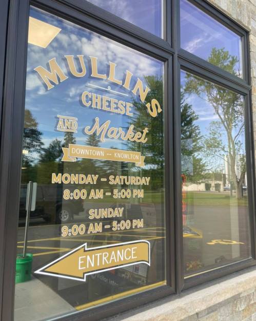 Sign on a window for Mullins Cheese Market, displaying hours and "Entrance" arrow.