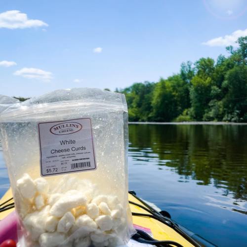 A bag of white cheese curds sits on a kayak, with a scenic river and trees in the background.