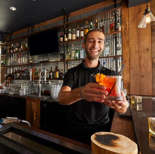 A smiling bartender holds a cocktail with an orange garnish, standing in a well-stocked bar.