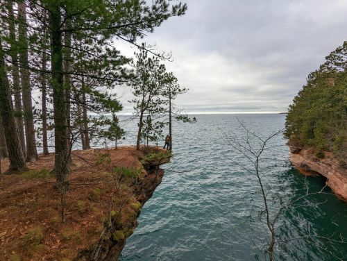 A person stands on a rocky cliff overlooking a calm lake surrounded by trees under a cloudy sky.