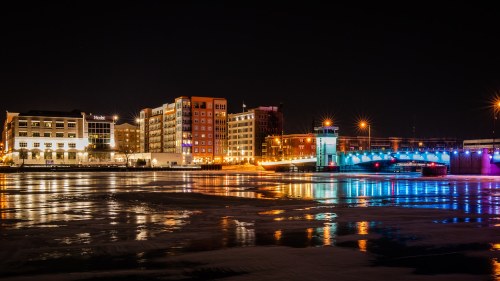 City skyline at night with illuminated buildings and a bridge reflecting on the water.
