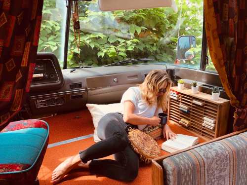 A person relaxes in a cozy van, reading a book and sipping coffee, surrounded by greenery.