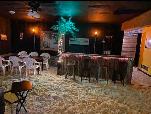 A cozy indoor space with sand flooring, a bar, chairs, and tropical decor, resembling a beach setting.