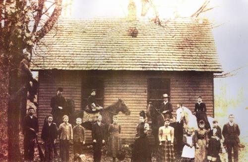 A historic group photo of a large family outside a wooden house, with some people on horseback, in a rural setting.