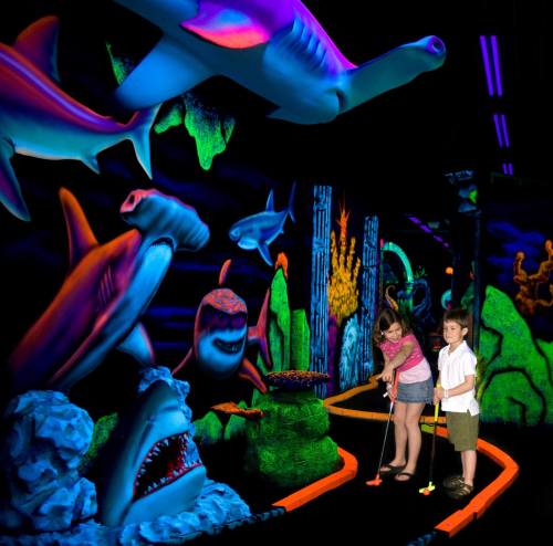 Two children play mini-golf in a vibrant, glow-in-the-dark underwater-themed course with colorful sea creatures.