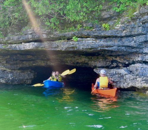 Two kayakers in colorful kayaks explore a rocky cave entrance on a sunny day, surrounded by green foliage.