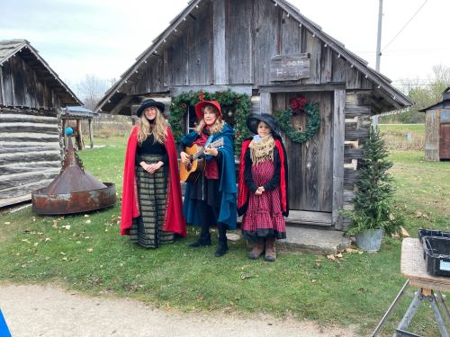 Three women in festive attire stand outside a rustic wooden cabin, one playing guitar, surrounded by greenery.